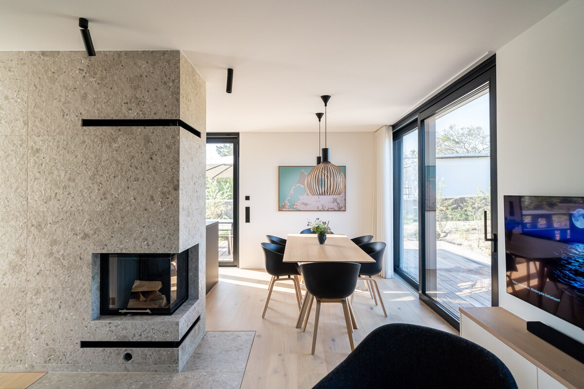 An open dining area features a wooden table surrounded by four black chairs. Large glass doors invite natural light and offer a view of the outdoor space. A modern fireplace is set within a textured stone wall, enhancing the contemporary design.
