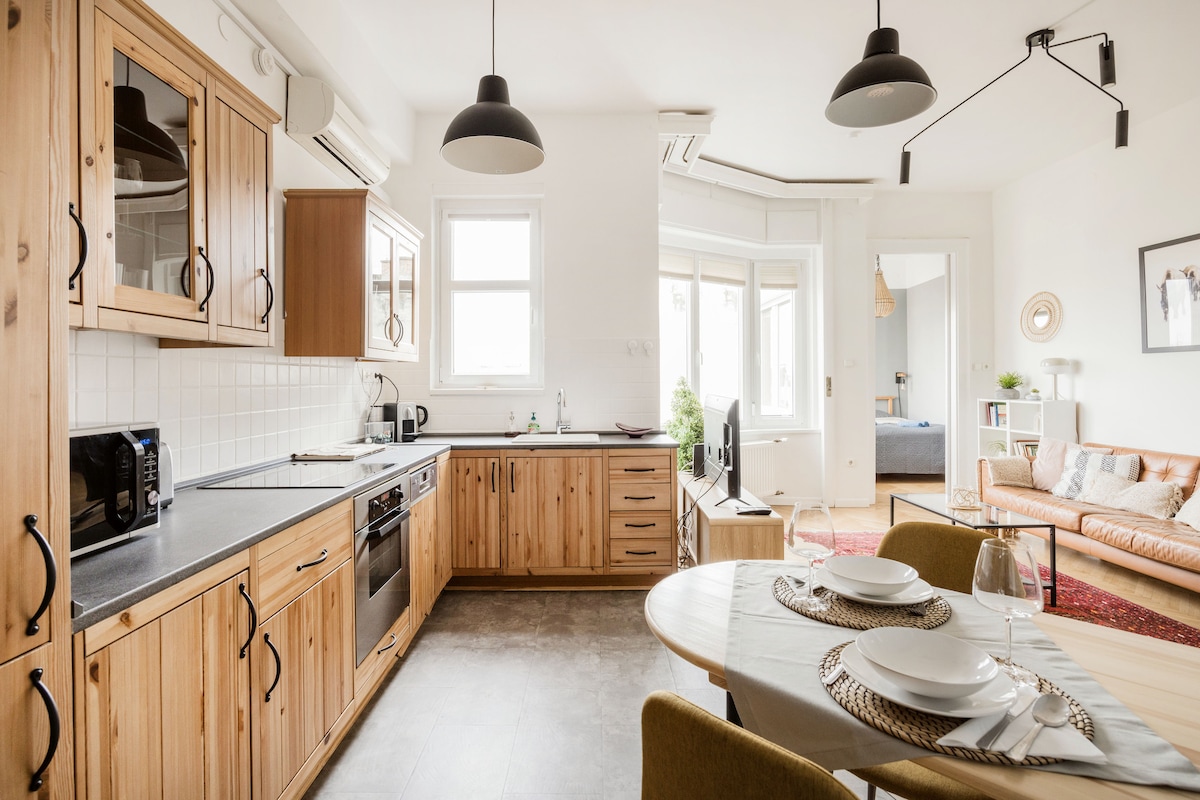 An open-plan kitchen and dining area feature wooden cabinetry and modern appliances. A round dining table is set with tableware, positioned near a cozy living room with a sofa. Natural light enters through a large window, creating an inviting space for socializing and cooking.