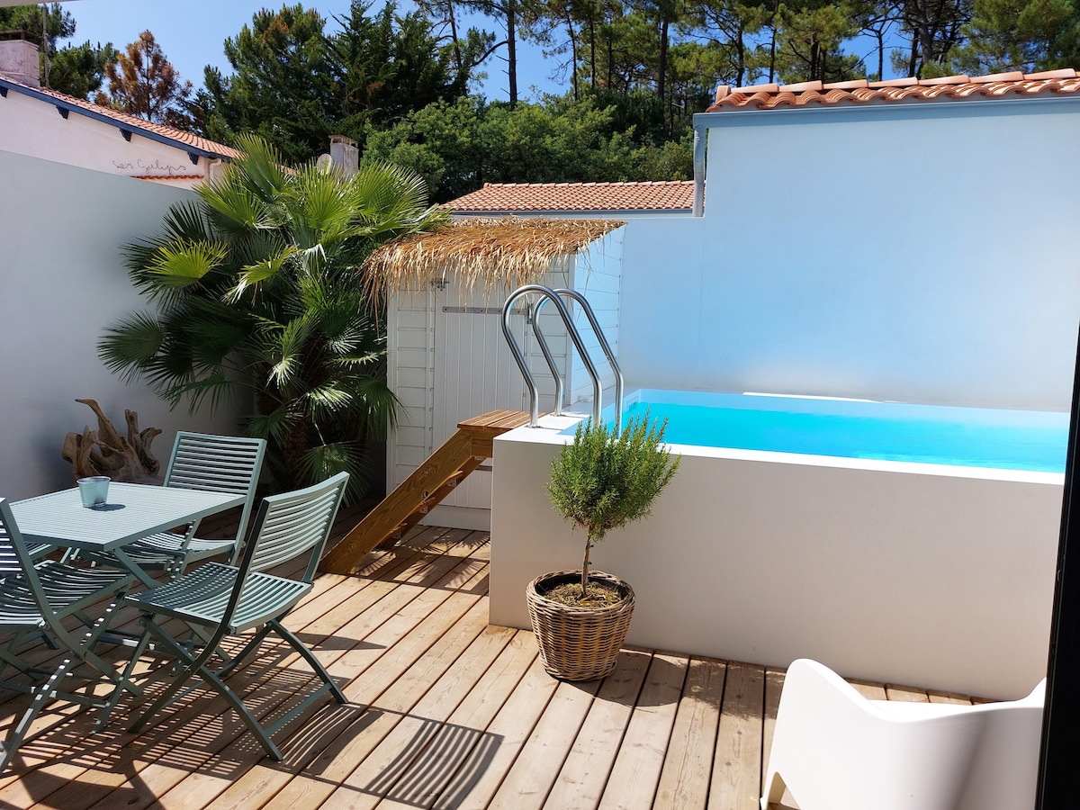 A private outdoor area features a small heated pool, flanked by wooden decking. A dining table with four chairs is positioned beside a potted plant, while a straw-covered structure provides shade. Lush greenery surrounding the space enhances the tranquil ambiance.