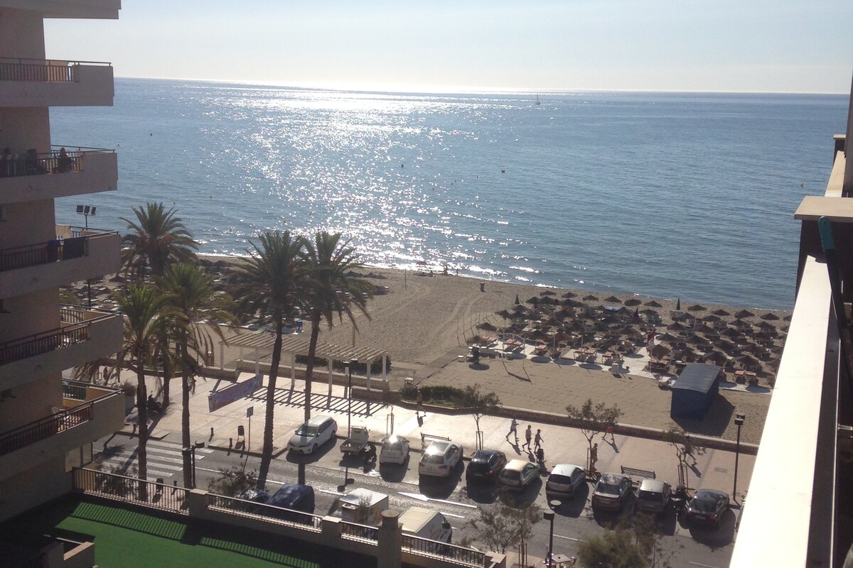 The image shows a serene seaside view with a sandy beach lined by palm trees. Sunlight reflects off the calm water, creating a glistening effect. Beach umbrellas and sun loungers are arranged on the sand, while a road filled with parked cars and pedestrians is visible in the foreground.