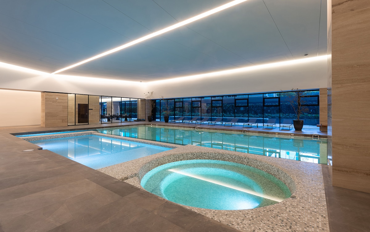An indoor pool area features a large swimming pool with a separate round hot tub, both with clear water. Large windows allow natural light to fill the space, highlighting the modern design and smooth tile surfaces. Lounge chairs are positioned around the poolside.