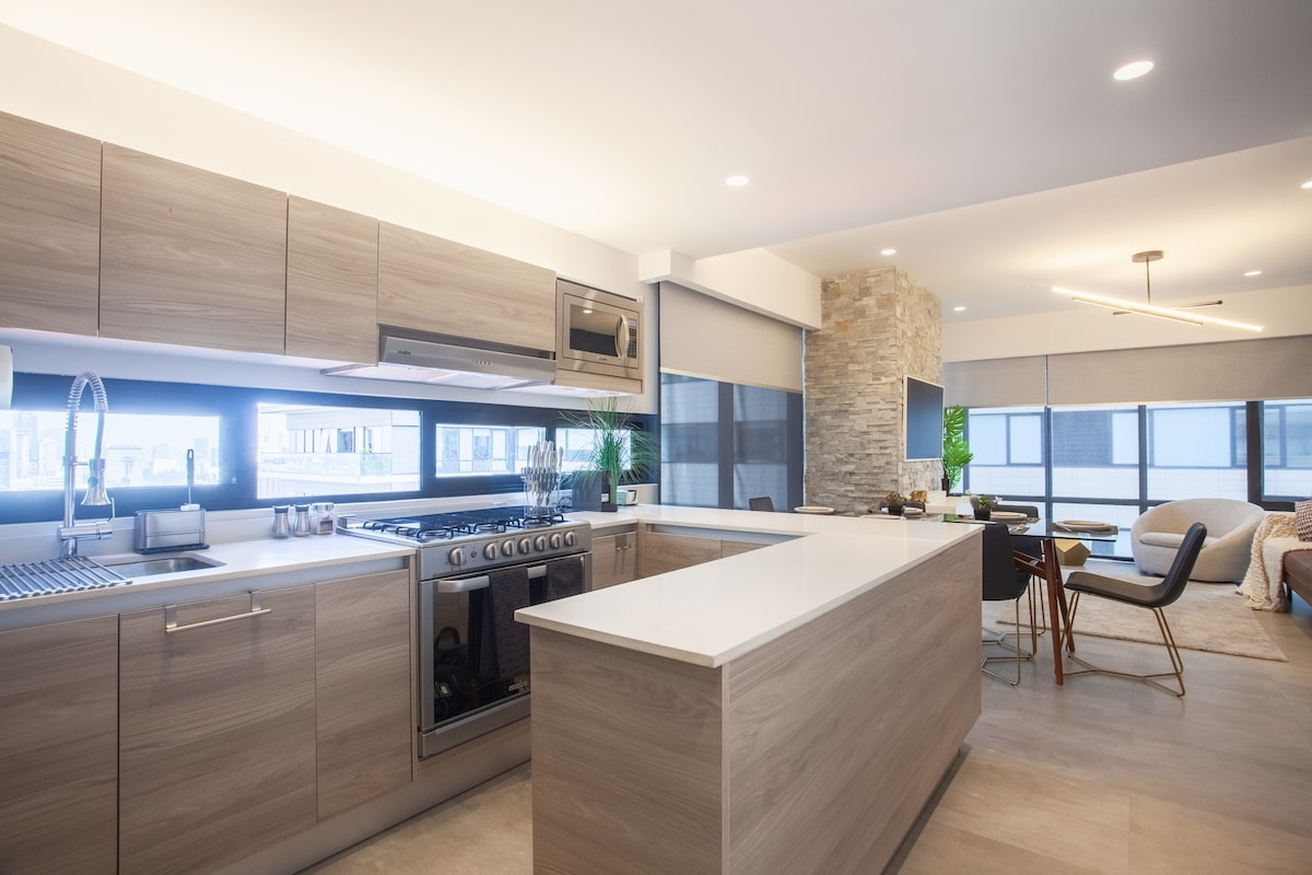 A spacious kitchen features modern cabinetry and a large island with a white countertop. High-end appliances, including a gas range, are visible. Natural light enters through large windows with roller shades, and a dining area is seen in the background, complemented by contemporary seating.