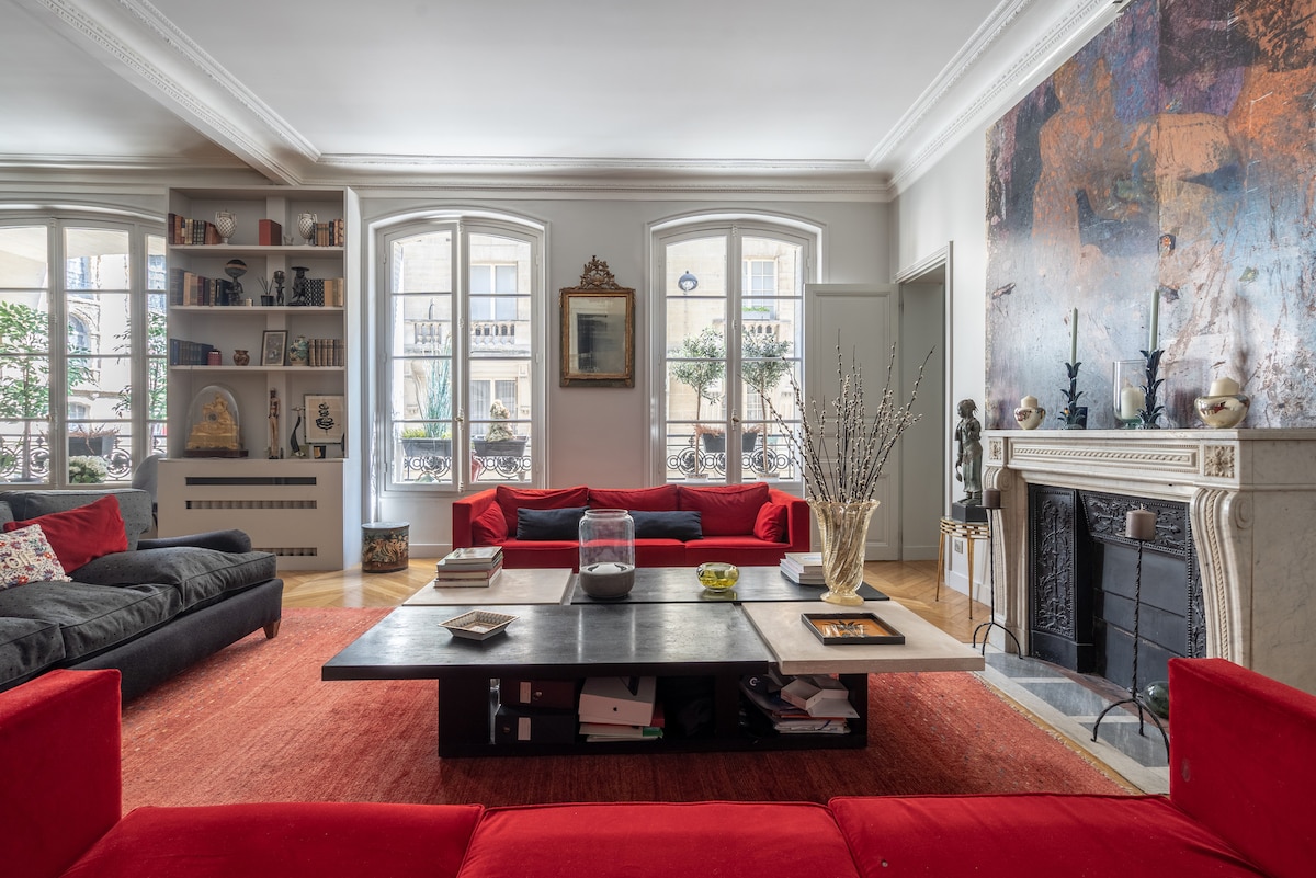 A spacious living room is highlighted by large windows allowing natural light to fill the space. Comfortable sofas in vibrant red and dark tones surround a central low table. A decorative wall features artwork, while bookshelves and a marble fireplace add character to the room.
