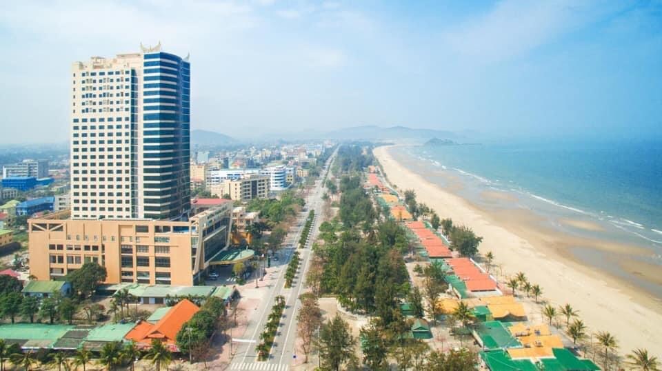An aerial view captures a beachfront area featuring a high-rise hotel and a long stretch of sandy beach. Lush palm trees line the coast, with colorful beach cabanas visible along the shore. A scenic promenade runs parallel to the beach, flanked by various buildings.