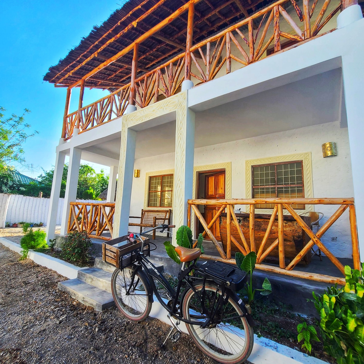 The exterior of the villa features a welcoming porch with wooden railings and seating. A bicycle is parked beside landscaped greenery, and the structure showcases a thatched roof accentuating its rustic design. Clear blue skies complement the inviting atmosphere.