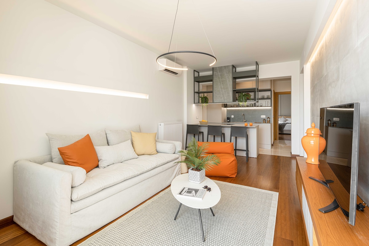 A light-filled living area features a neutral-colored sofa adorned with orange cushions. A round coffee table sits on a textured rug, complemented by a small potted plant. The open-concept kitchen is visible in the background, showcasing modern cabinetry and appliances.
