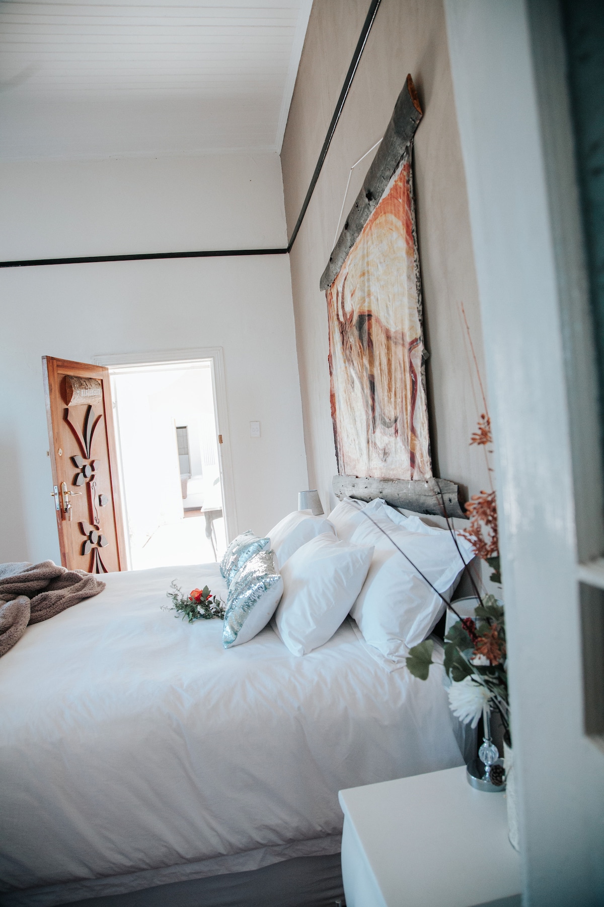 A serene bedroom is showcased, featuring a spacious bed adorned with white linens and decorative pillows. A vibrant artwork hangs on the wall, while a bedside table holds a small arrangement of flowers. A door opens to a sunny outdoor area, enhancing the room's brightness.