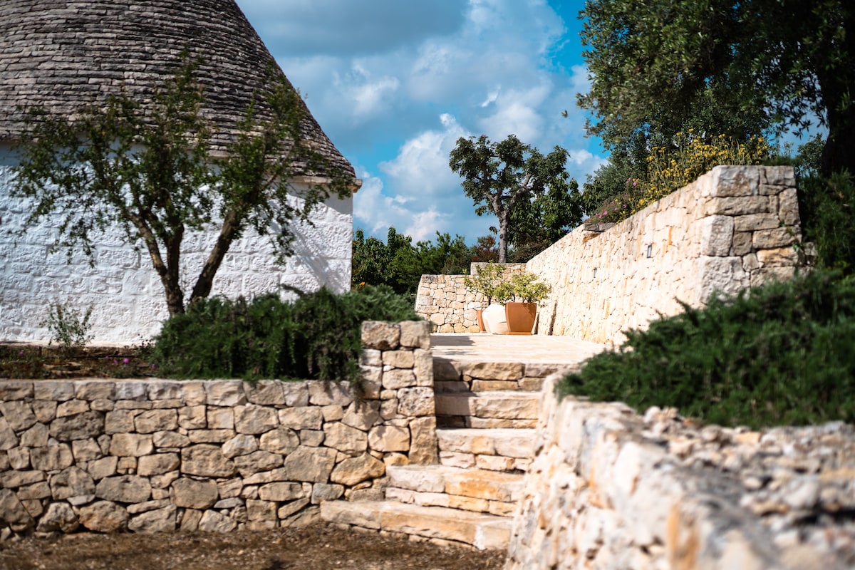The image captures a serene outdoor pathway leading to a traditional trullo structure. Natural stone walls frame the path, which is lined with greenery and flowering plants. The scene is enhanced by a bright sky scattered with clouds, inviting a peaceful atmosphere.