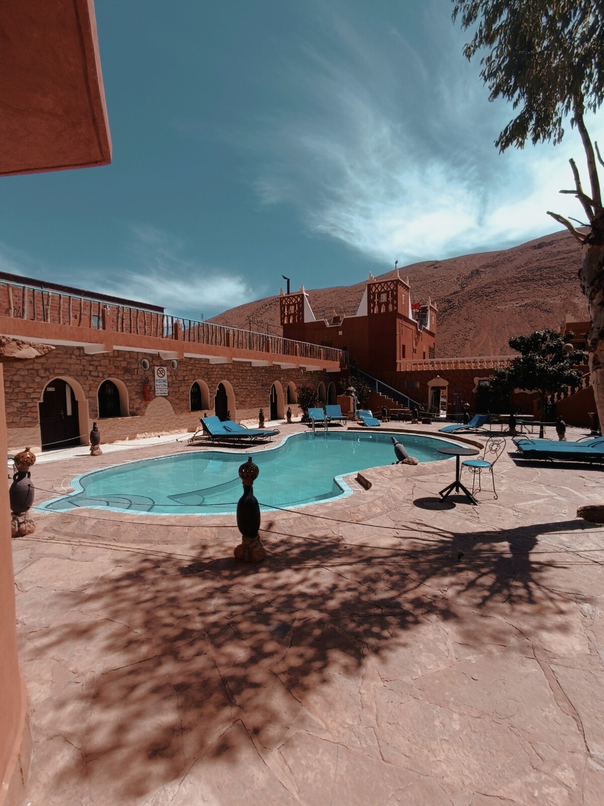 An outdoor area features a swimming pool surrounded by lounge chairs. The stone structure outlines the poolside, while the backdrop showcases rolling hills and a vibrant sky. Shadows are cast on the stone surface, adding depth to the tranquil environment.
