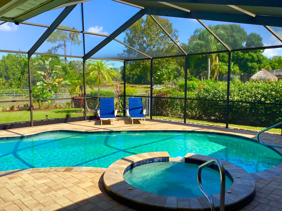 A screened-in tropical pool area features a refreshing pool with a separate spa. Two loungers are positioned beside the water, while lush greenery surrounds the space, providing a serene backdrop. Sunlight filters through the mesh, creating a bright and inviting atmosphere.