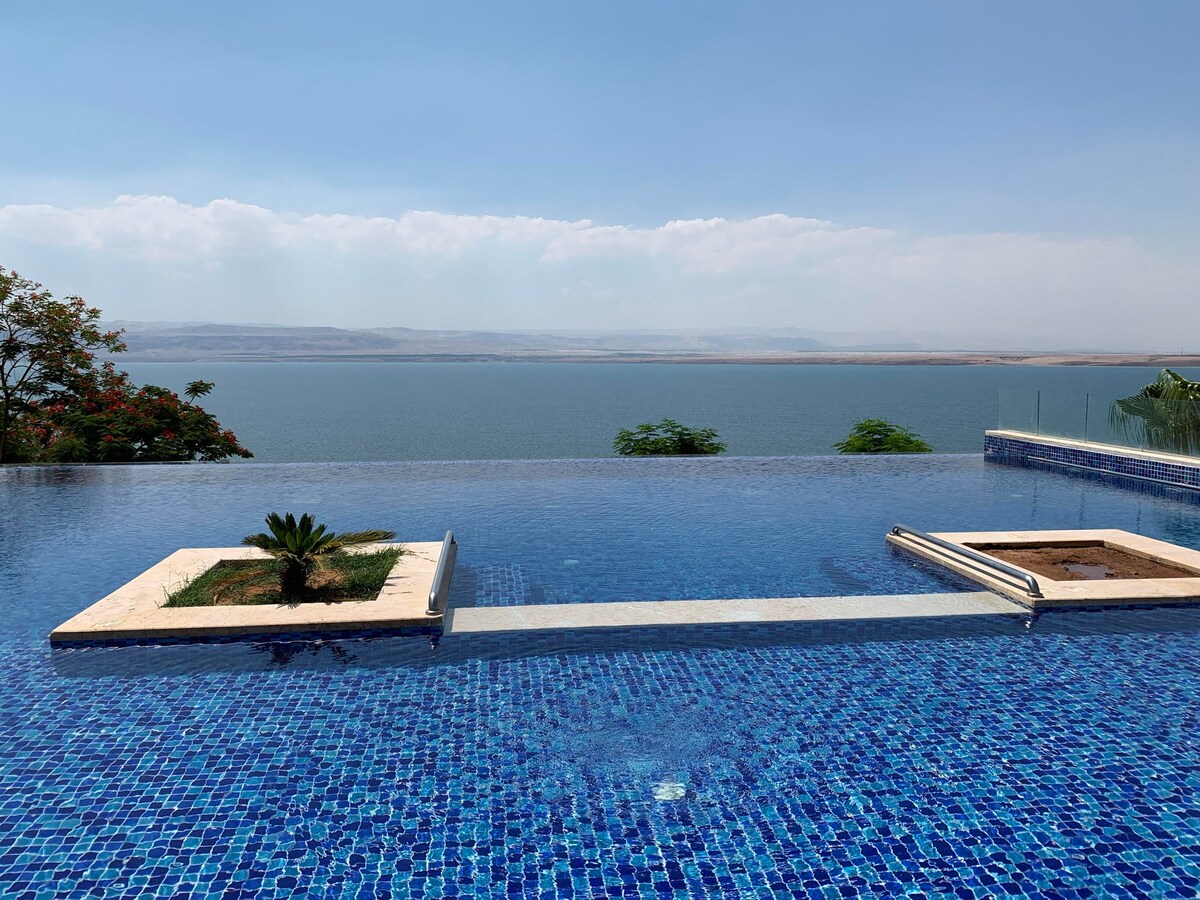 An infinity pool stretches toward a horizon, featuring mosaic tiles in shades of blue. Lush greenery frames the pool area, while a serene water body extends in the background under a clear sky, providing a peaceful view.