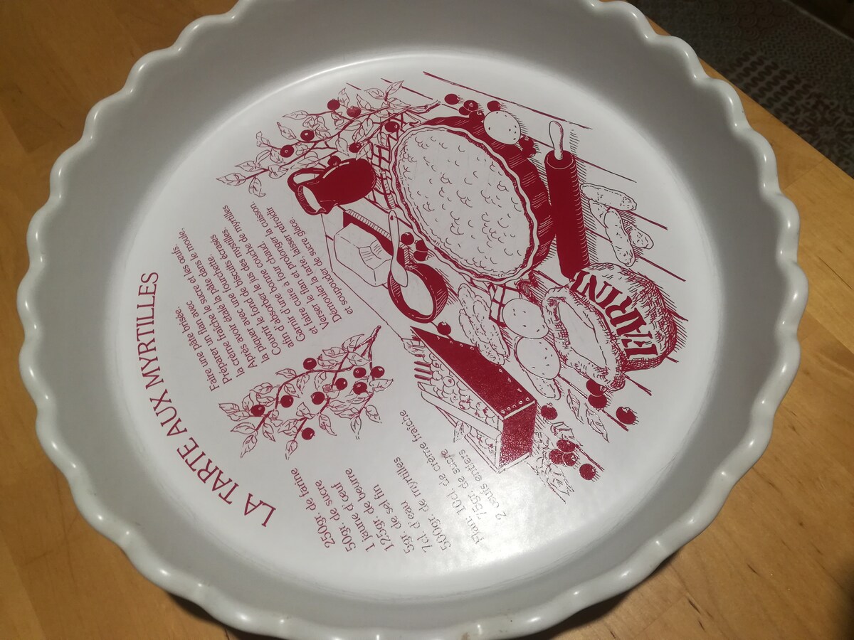 A decorative pie dish features a red illustration depicting a blueberry pie and related ingredients. Text describing a recipe is printed in a circular fashion around the edge, with a ruffled lip providing an elegant touch.