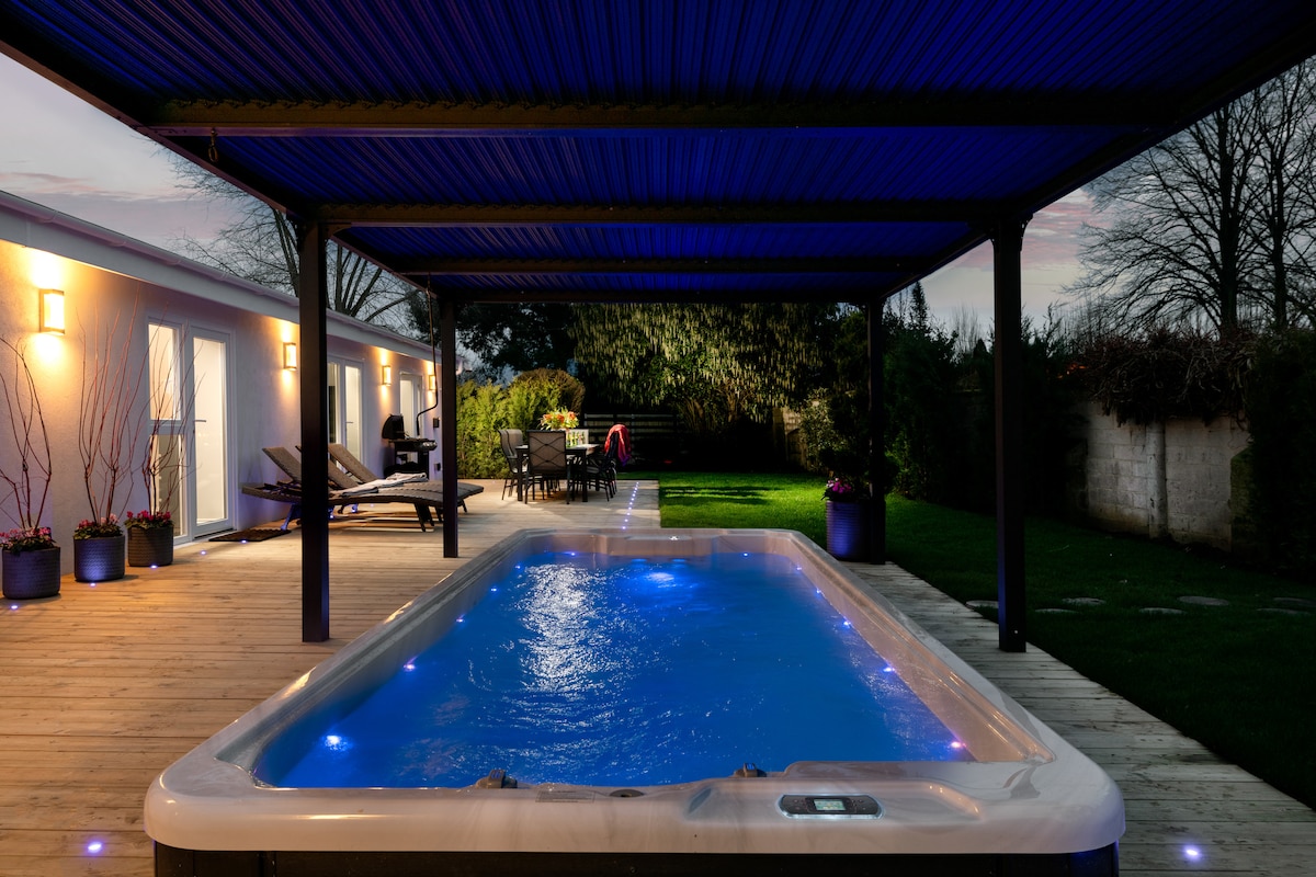 A private spa area features a well-lit hot tub with bubbling water, under a sheltered pergola. Surrounding it are wooden decking and plush lounge chairs. The green garden is visible beyond, providing a tranquil setting, with dim lighting enhancing the evening ambiance.