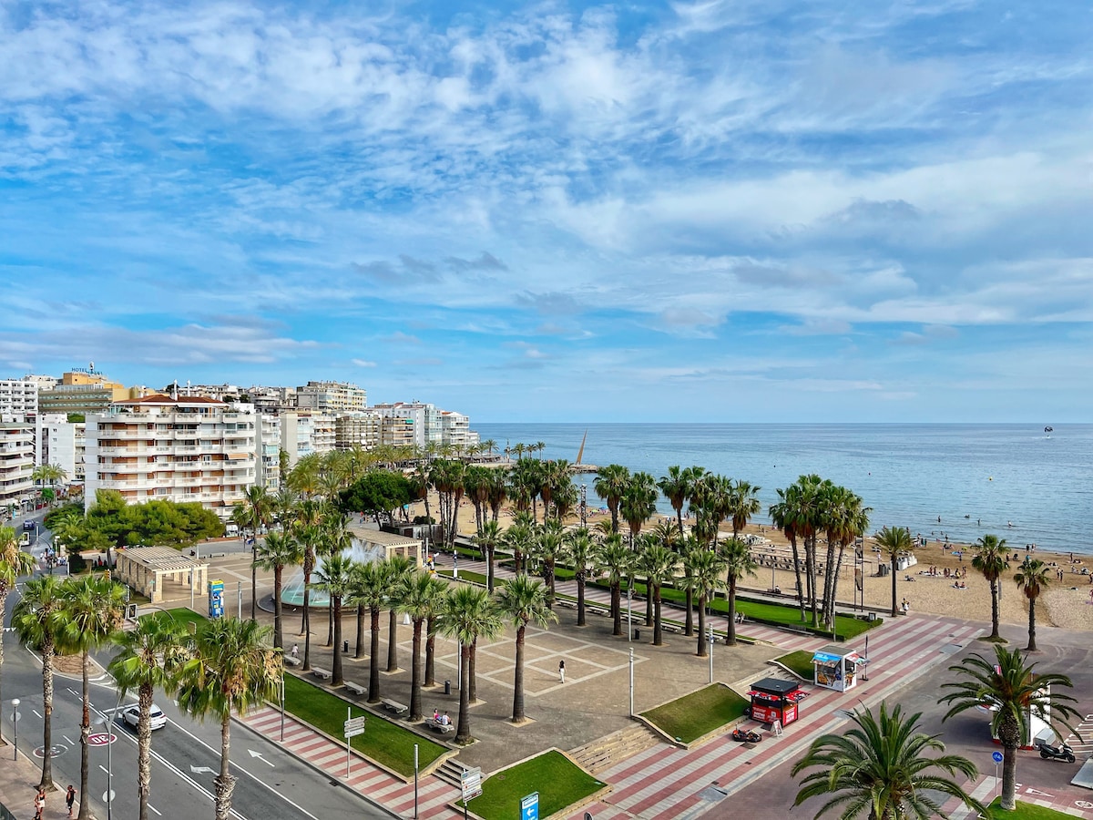 A view of the vibrant beachfront area showcases a line of palm trees accompanied by a spacious park. Calm waters can be seen in the distance, with sandy shores just beyond. Buildings are visible along the coastline, contributing to the lively coastal atmosphere.