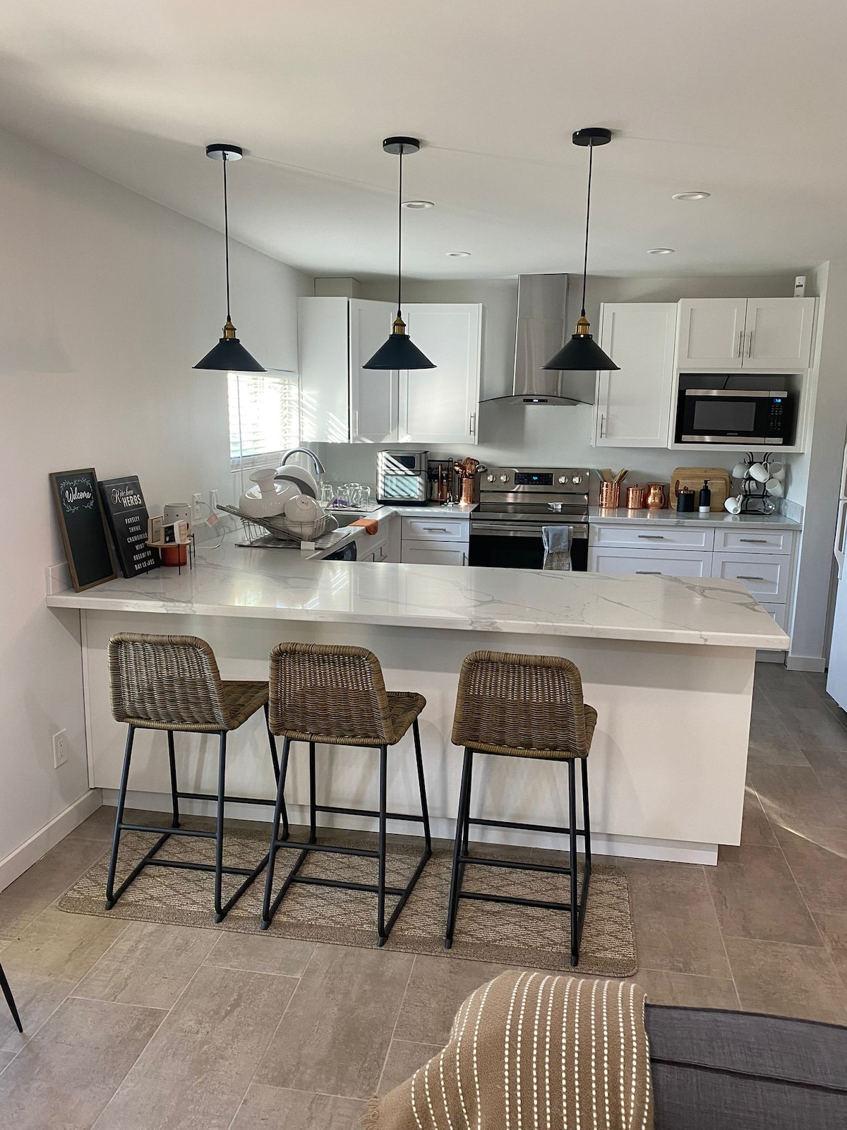 A modern kitchen features sleek white cabinetry and stainless steel appliances. Pendant lights hang above a bar with three woven stools. A spacious countertop offers ample prep space, complemented by a stylish backsplash and well-organized kitchen essentials in the background.