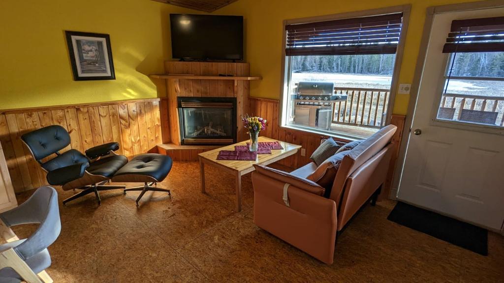 A warm living area features a modern brown sofa and a cozy chair, positioned near a fireplace. A flat-screen TV is mounted above a wooden mantel. Natural light enters through a window, showcasing views of the surroundings, and a door leads outside.