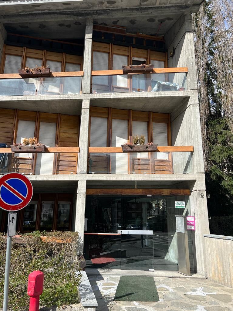 The exterior of a multi-story building showcases a modern design with wooden accents and glass railings. Balconies with planter boxes are visible, and a sign indicating guest access is positioned near the entrance. Surrounding greenery adds a natural touch to the environment.