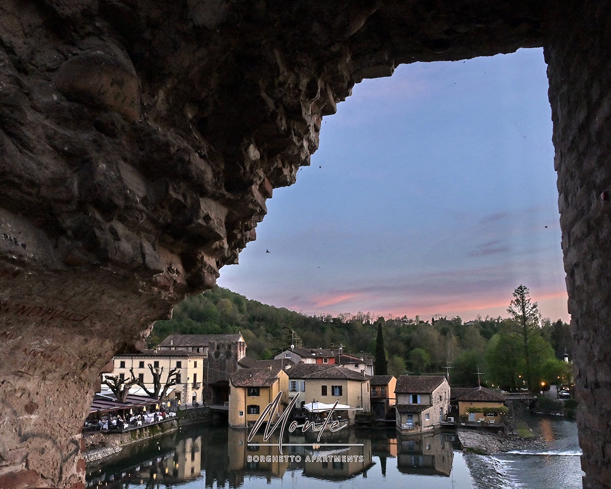 A scenic view is captured through an archway, revealing charming buildings along a tranquil waterway. Lush green trees frame the landscape, while soft colors of dusk create a serene backdrop. The historical stone structure adds texture to the image, providing a glimpse of local architecture.