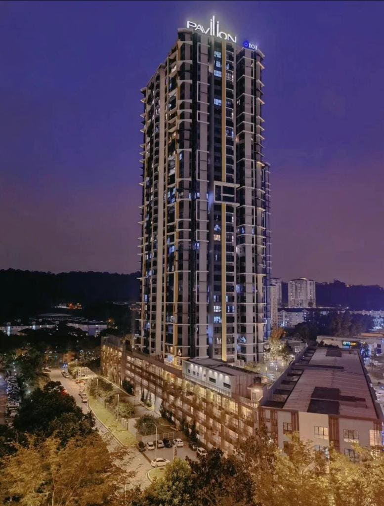 An impressive tall building is captured at dusk, featuring a well-lit façade that highlights its modern architectural design. Surrounding greenery is visible, along with a softly illuminated urban landscape below, creating a serene evening atmosphere.