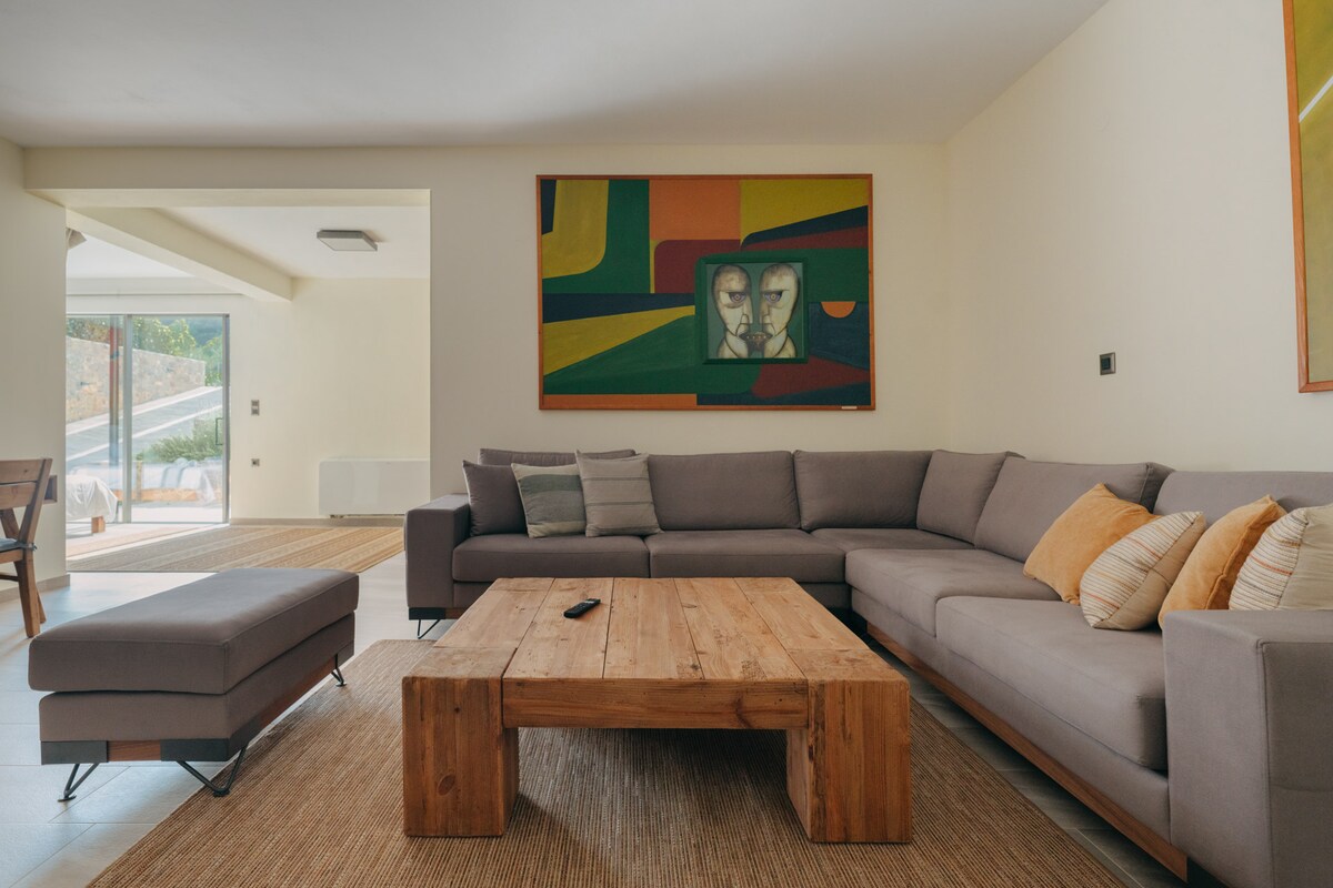 A spacious living area features a large, L-shaped gray sofa complemented by wooden accents and decorative cushions. A rustic wooden coffee table sits at the center, while a colorful wall art piece adds a vibrant touch. Natural light floods the space from nearby glass doors.
