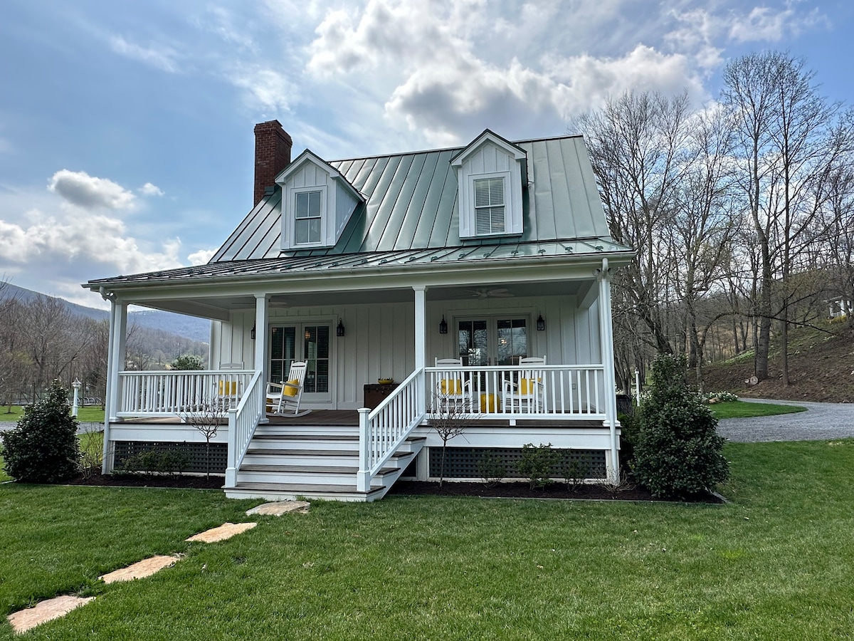 A charming white cottage with an inviting front porch is depicted. Yellow chairs are positioned on the porch, enhancing the welcoming appearance. Lush green grass surrounds the home, with trees in the background, creating a serene outdoor setting.