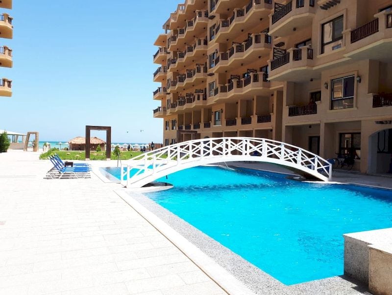 A wide pool is situated in the foreground, featuring a small white bridge that spans across it. Surrounding the pool, sun loungers are arranged on the tile deck. The apartment building's balconies overlook the pool area, with views of the beach and sea visible in the background.