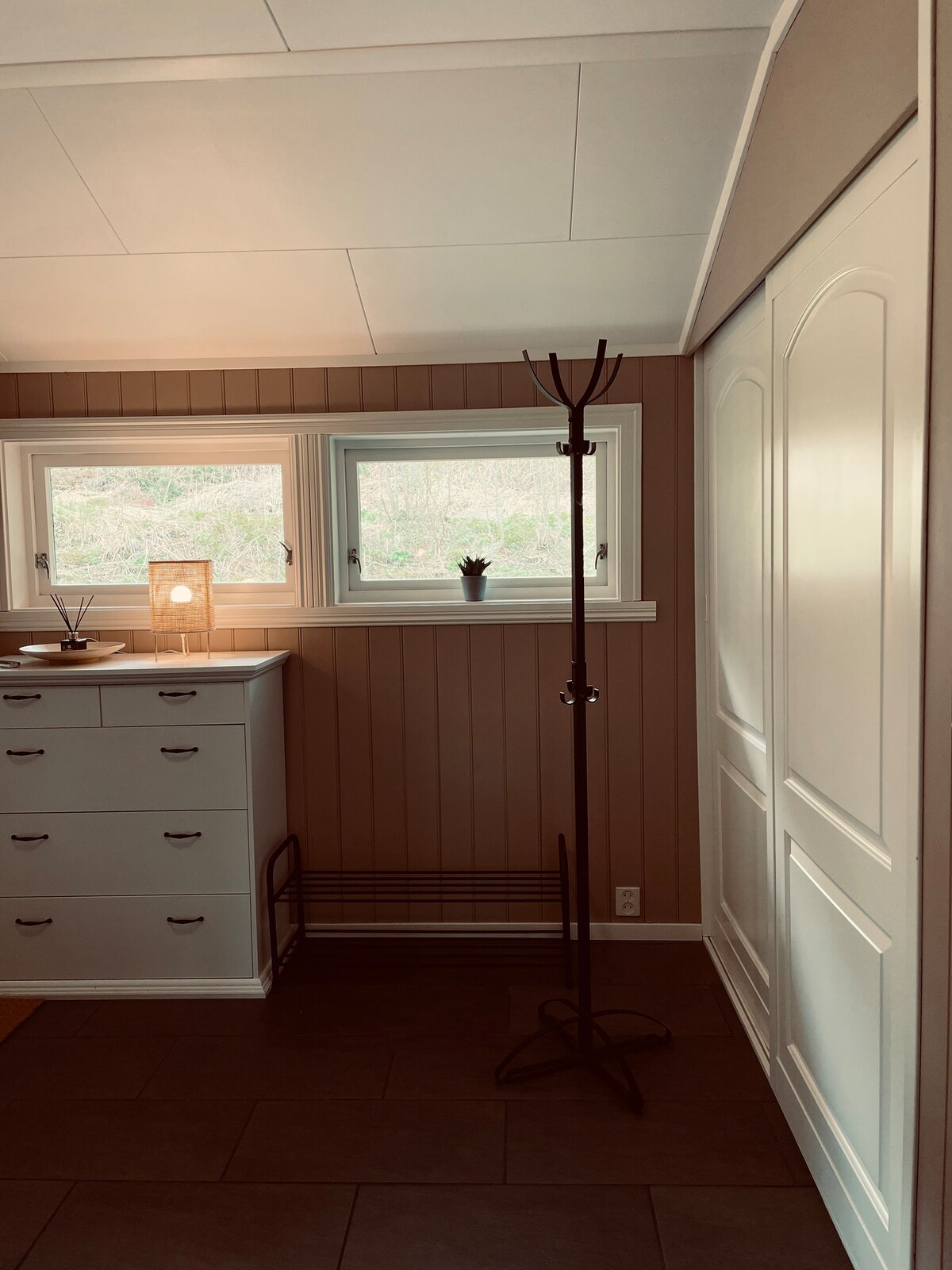 A light-filled entryway features two windows with greenery visible outside. A minimalist coat rack stands against the wall next to a white dresser with multiple drawers. Natural tones of the walls create a warm and welcoming space.