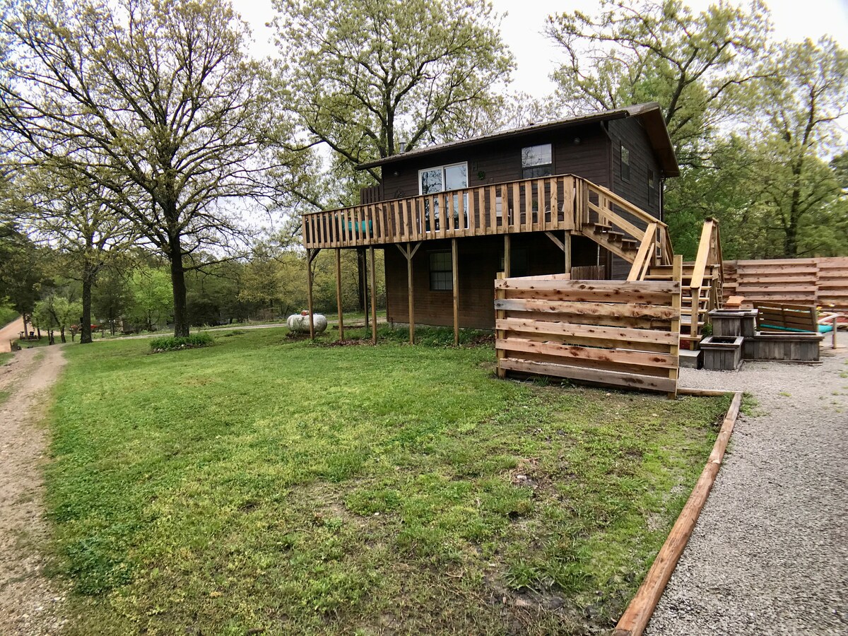 A two-story cabin is positioned on a grassy area, surrounded by trees. It features a wooden deck with steps leading up to the entrance. The exterior is clad in dark wood, while a gravel path provides access to the property, showcasing a serene country setting.
