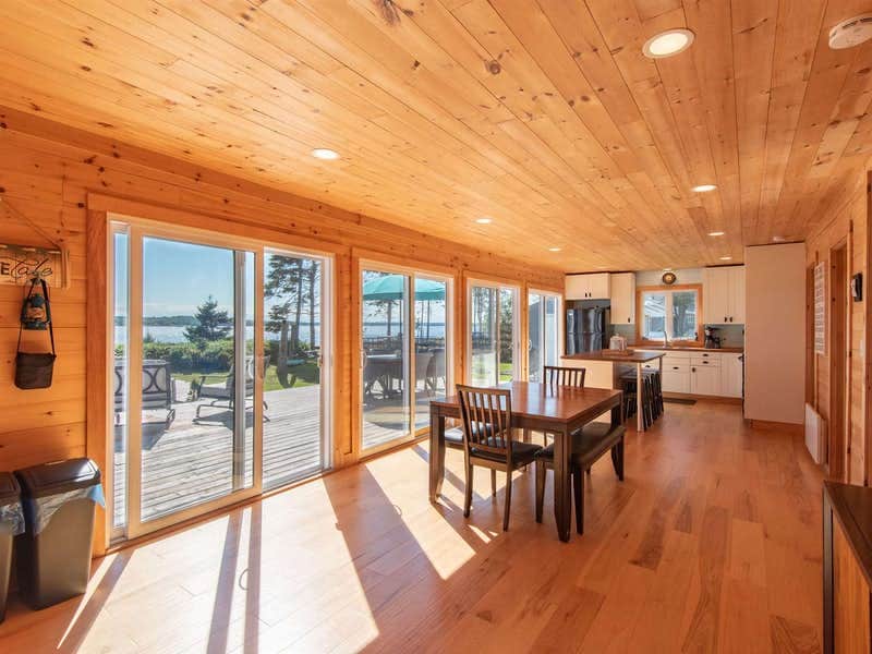 A spacious interior area features warm wooden walls and a wooden ceiling. Large sliding glass doors provide access to a deck with outdoor seating, allowing natural light to fill the space. A dining table is positioned beside the kitchen, enhancing the open and inviting layout.