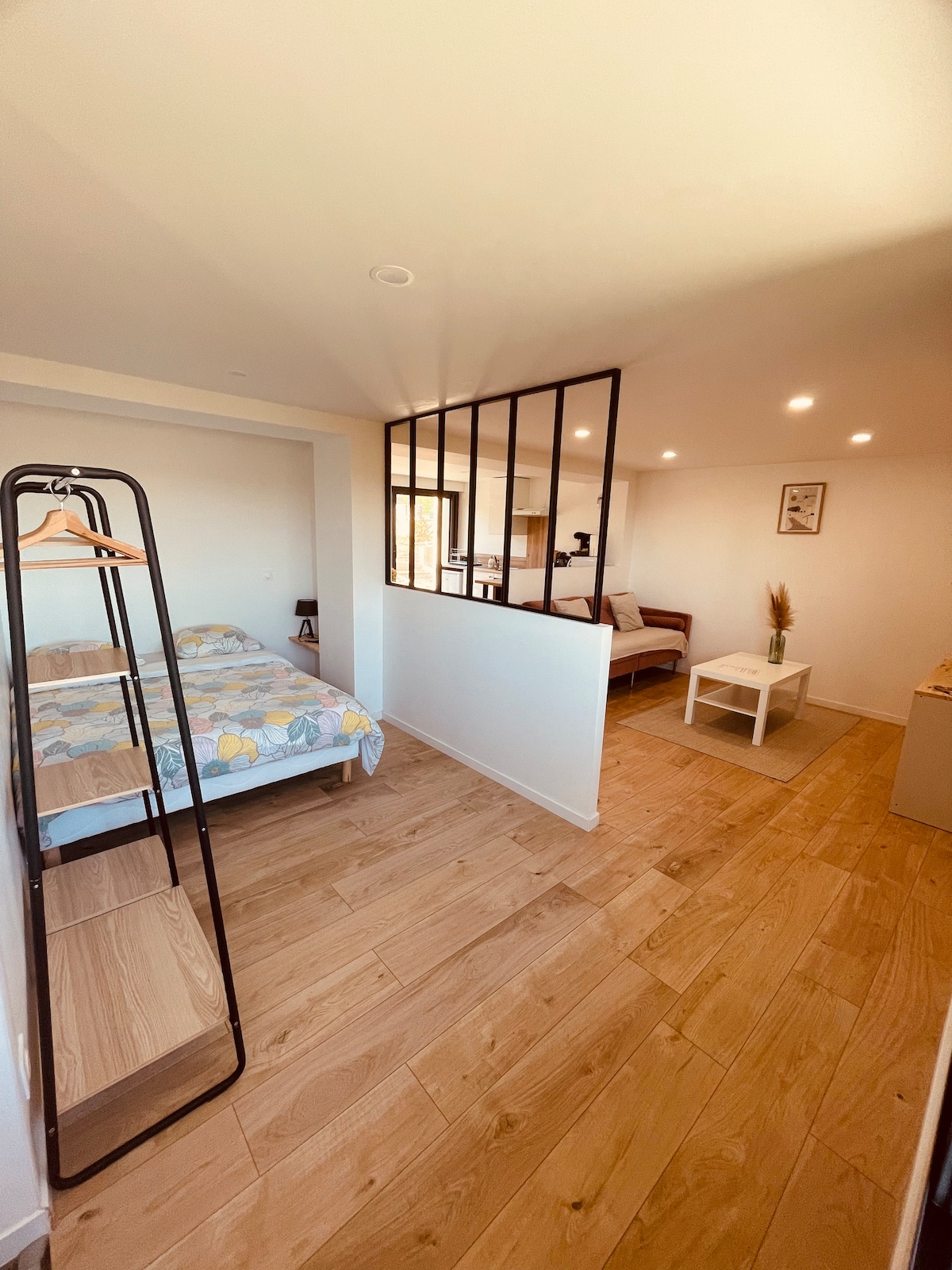 A well-lit studio apartment features a spacious sleeping area with a patterned bedspread. A decorative partition separates the sleeping space from a small sitting area, which includes a couch and a coffee table. Warm wood flooring contributes to the inviting atmosphere.