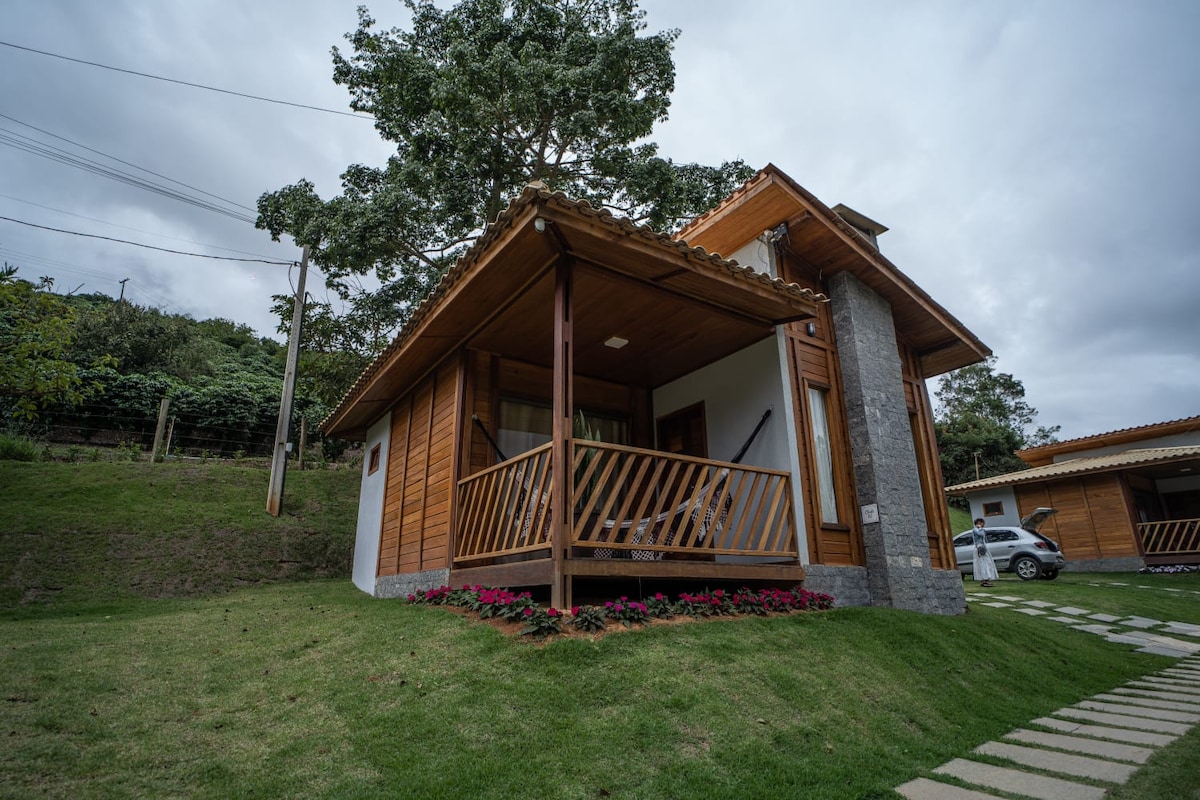 A charming wooden chalet is set against a green hillside. A covered porch with seating faces a neatly landscaped area, enhancing the homely atmosphere. The exterior features natural stone accents and is surrounded by colorful flowers and neatly arranged stepping stones.