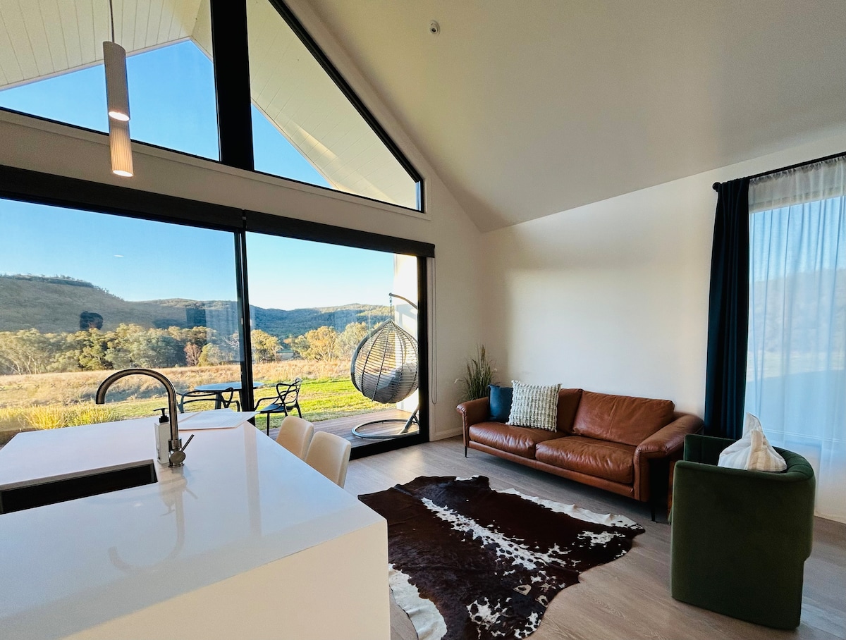 A spacious living area features large windows that frame expansive views of rolling hills. A brown leather sofa is positioned alongside a cozy green armchair, and a modern hanging chair adds a stylish touch. Natural light brightens the space, complementing the neutral decor.