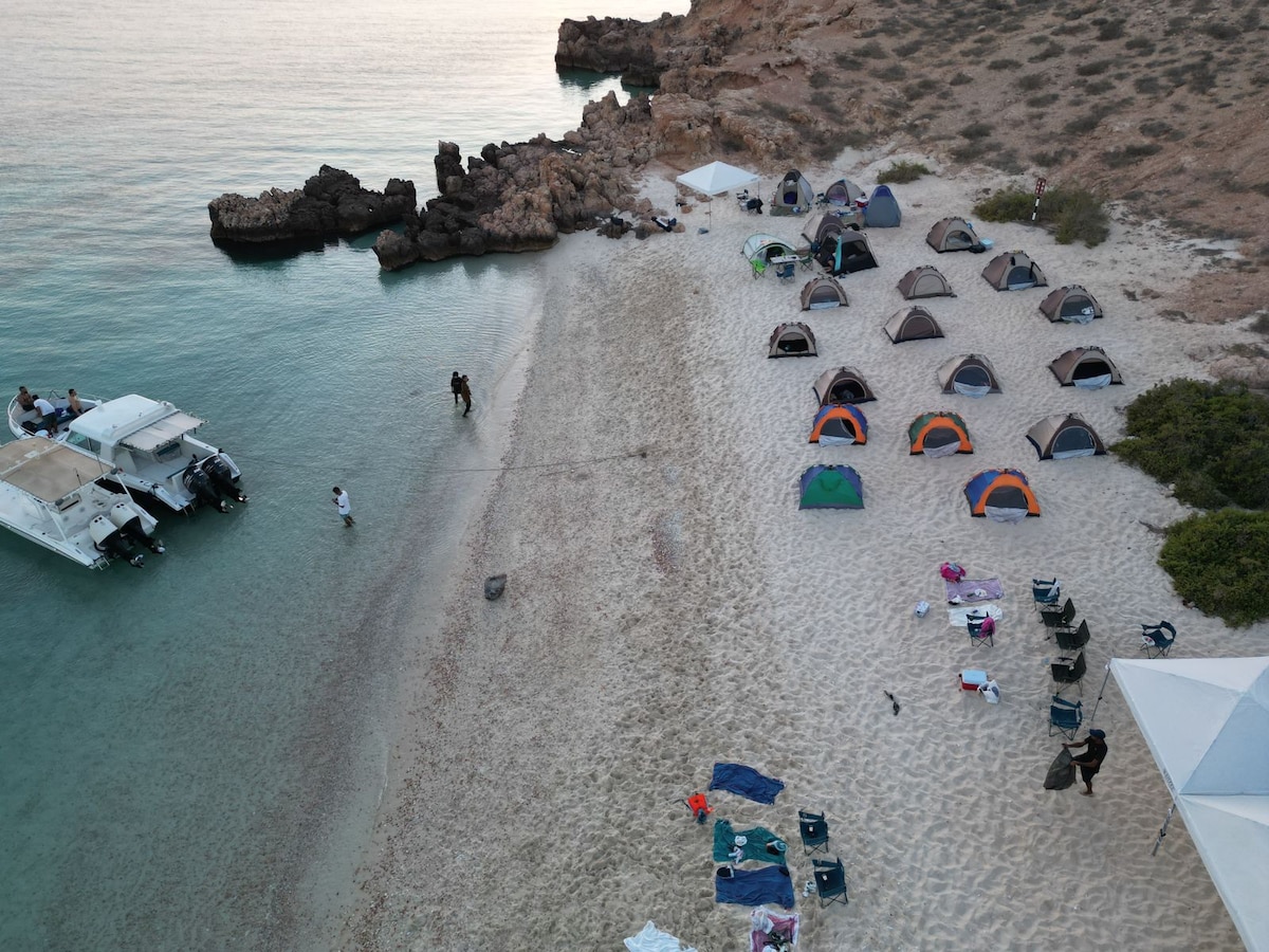 A sandy beach is bordered by clear waters and rocky formations. Several tents are pitched on the shore, complemented by lounging chairs and scattered gear. Two boats are visible near the shoreline, enhancing the coastal setting.
