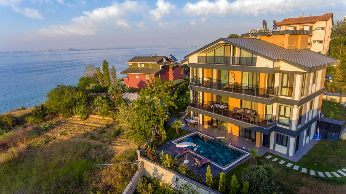 A modern villa overlooking the sea, featuring multiple levels and large glass windows that offer stunning views. An expansive pool is situated in the backyard, surrounded by greenery, enhancing the outdoor spaces. The property’s design merges seamlessly with the natural landscape.