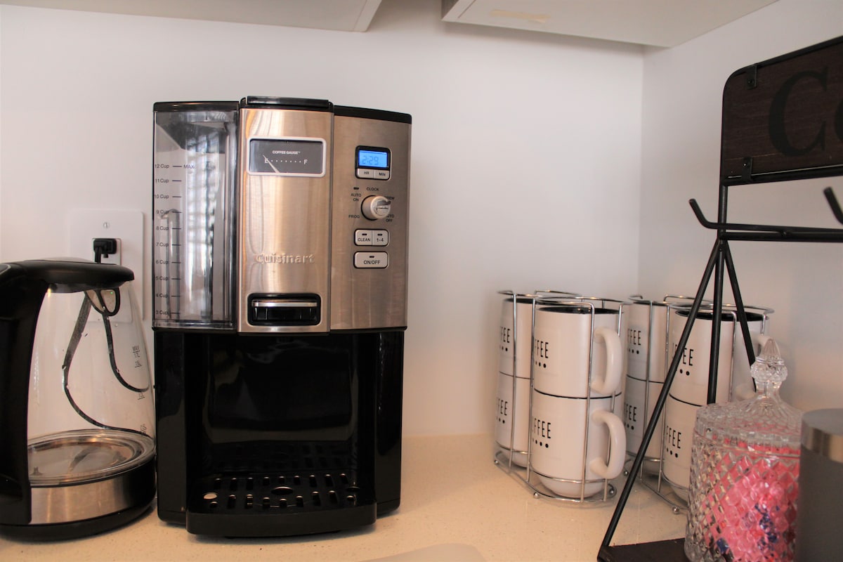 A modern coffee maker is showcased on a light countertop, accompanied by a silver kettle and neatly arranged mugs labeled 'COFFEE.' Light reflections can be seen on the surface of the coffee maker, while a decorative item adds a pop of color nearby.