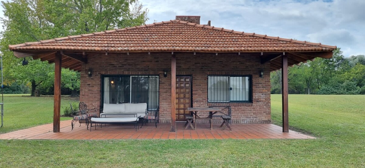The exterior of a single-story brick house is shown, featuring a gabled tiled roof and a spacious covered porch. A seating area with a sofa and a table is visible, surrounded by expansive green lawns and trees, creating a serene outdoor environment.