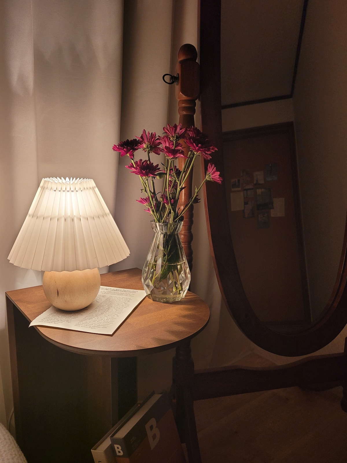 A bedside table is presented, featuring a soft-lit lamp with a pleated shade and a vase of purple flowers. The surface hosts an open book, and a full-length mirror is positioned nearby, reflecting the warm ambiance of the space.