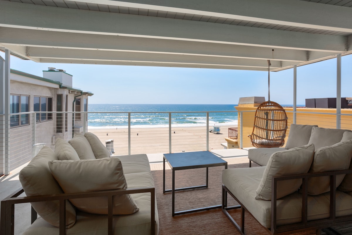An outdoor seating area is featured, overlooking the sandy beach and ocean. The space includes a light-colored cushioned seating arrangement and a small table. A modern hanging chair is also visible, creating a relaxed atmosphere. The expansive ocean view is unobstructed.