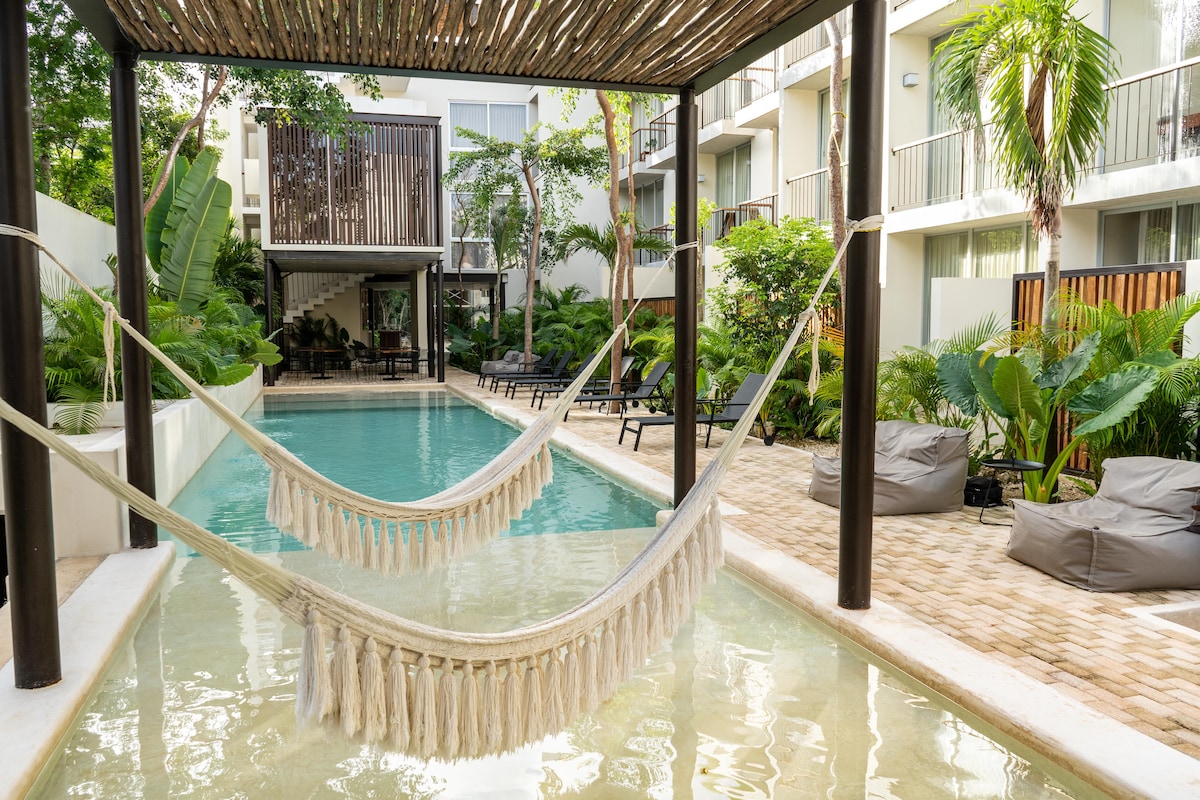 A serene outdoor area features two hammocks strung over a calm pool, surrounded by lush greenery and tropical plants. Comfortable bean bag chairs are positioned by the clear water, while a modern building with balconies is partially visible in the background.