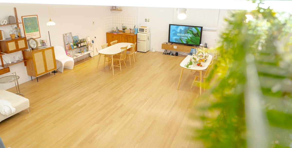 An open studio space features light wood flooring and modern furnishings. A large TV is positioned on one side, while tables are arranged for dining and activities. Several chairs surround the tables, and a small kitchenette can be seen in the background.