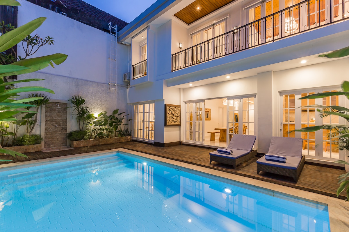 A clear view of the villa's serene pool area is presented, featuring two sun loungers positioned by the water's edge. The spacious outdoor deck is surrounded by tropical plants, while the villa’s modern, bright design is showcased through large glass doors leading to the interior.