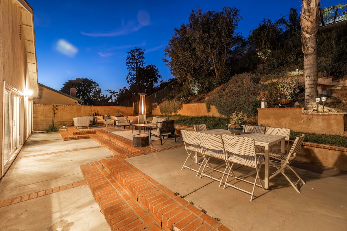 An outdoor space is showcased with a large dining table surrounded by white chairs. Comfortable lounge seating is arranged near a fire pit, and soft lighting creates a relaxing atmosphere. The backdrop features terraced greenery and a gently sloping hillside.