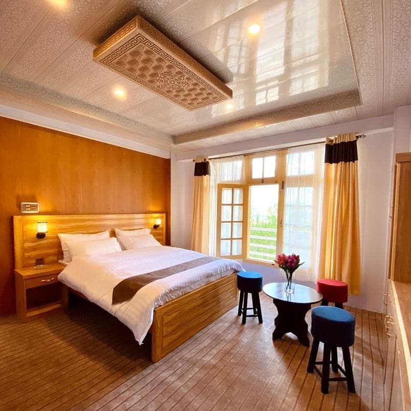A cozy bedroom features a spacious bed with a light-colored duvet. Natural light enters through large windows, complemented by soft curtains. Colorful stools and a round table create a warm seating area. The ceiling is intricately designed, adding a unique touch to the room.