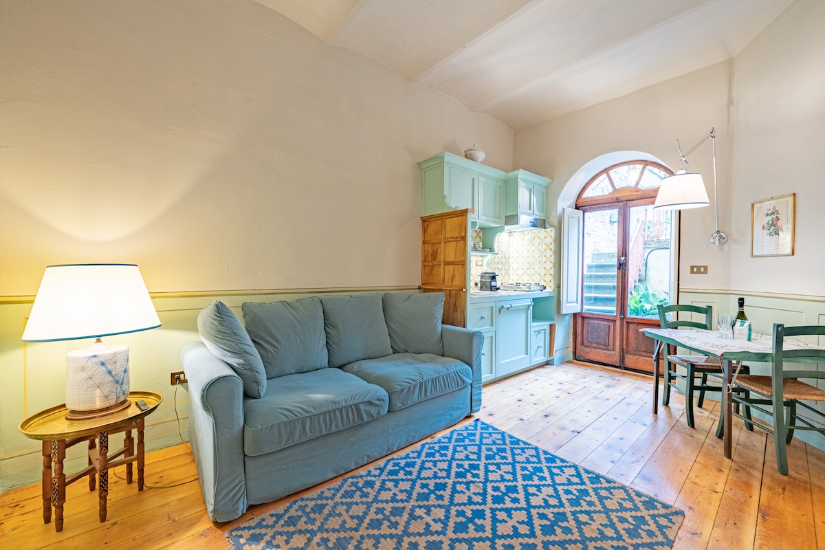 The living area features a comfortable blue sofa, warm wooden flooring, and an inviting dining table. Light enters through the arched window, highlighting the vintage-style kitchen equipped with teal cabinetry and patterned backsplash. A simple lamp adds soft illumination to the space.
