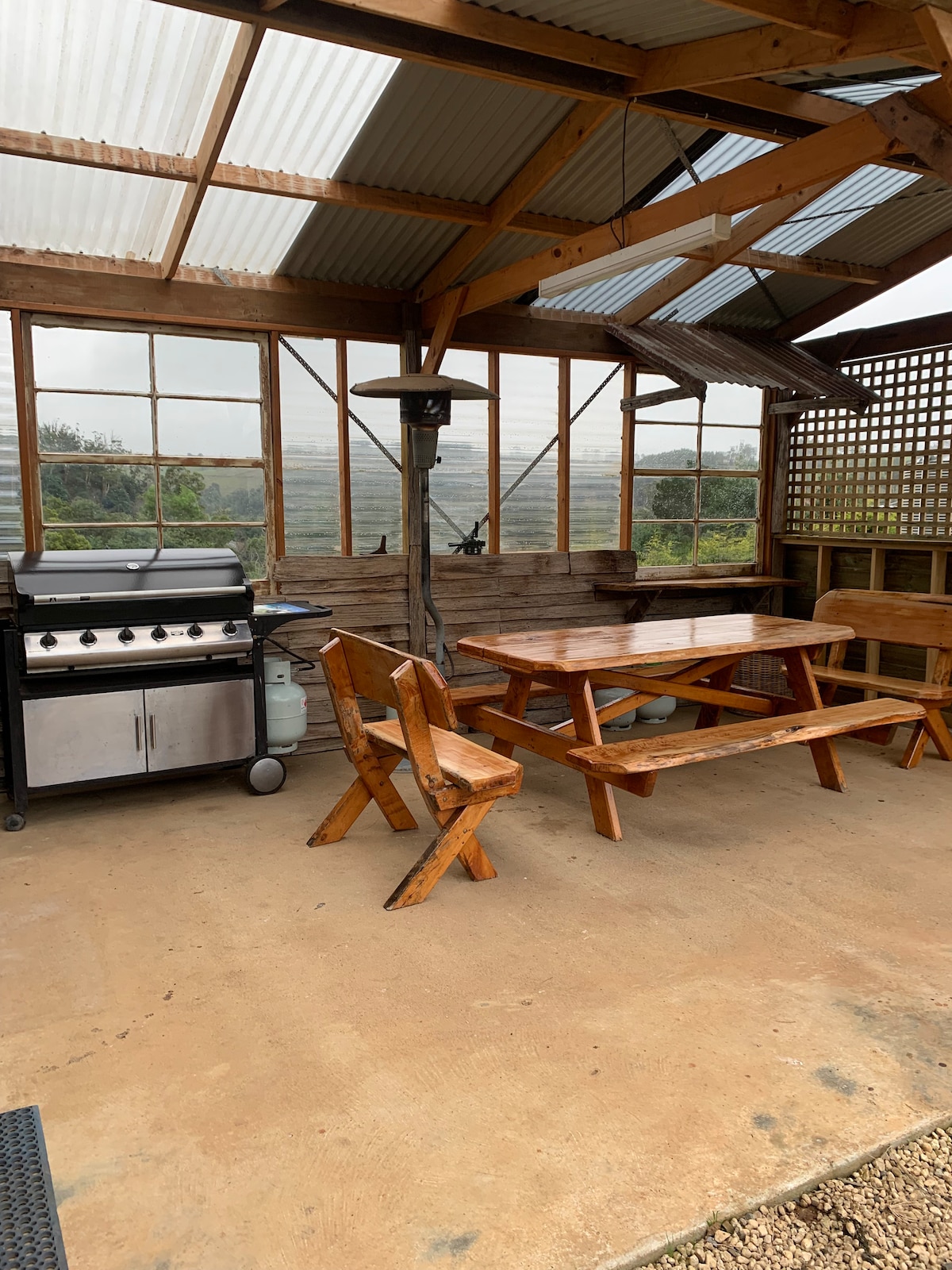 An outdoor dining area is presented with a wooden table and benches beneath a roofed structure. A grill is positioned to one side, and large windows provide a view of the surrounding landscape. The space offers a relaxed atmosphere for dining al fresco.