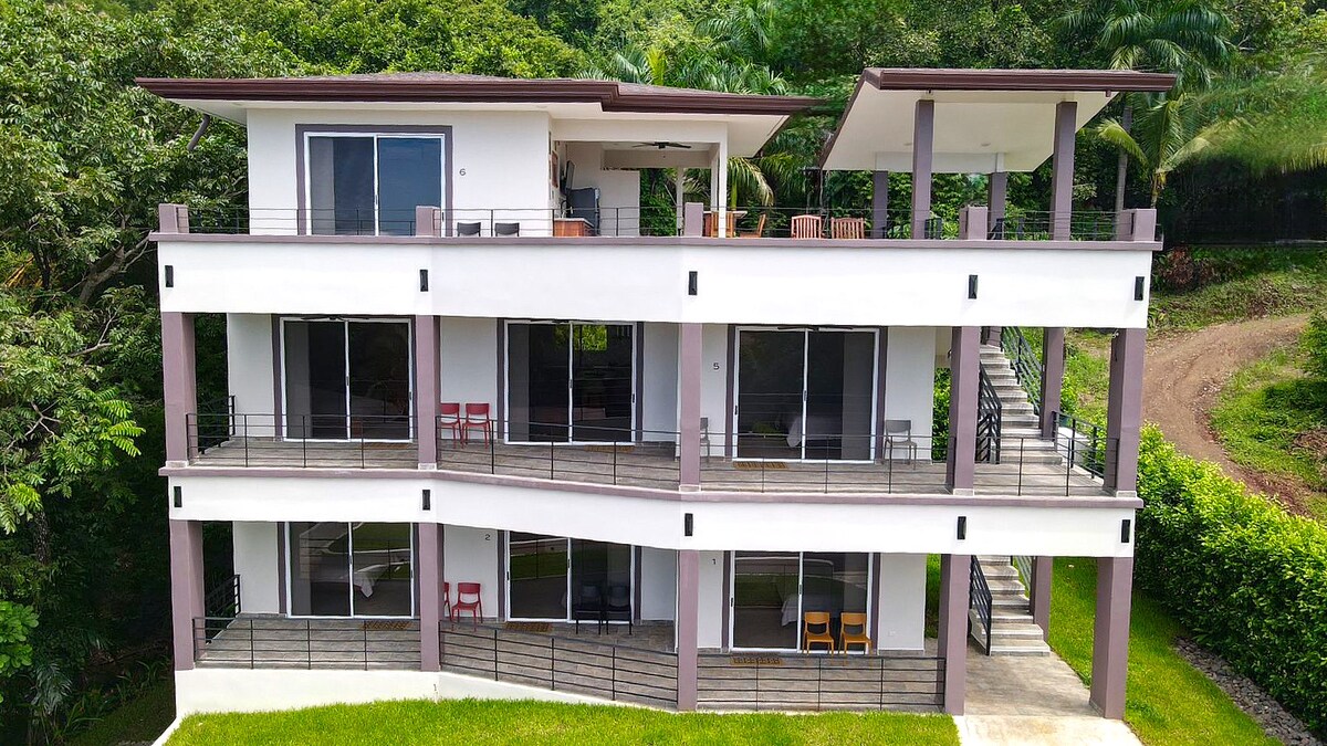 A contemporary multi-level building is surrounded by lush greenery, showcasing large glass doors and windows that offer expansive views. The structure features spacious patios furnished with simple seating arrangements. A sloping pathway leads up the building, enhancing the natural landscape.