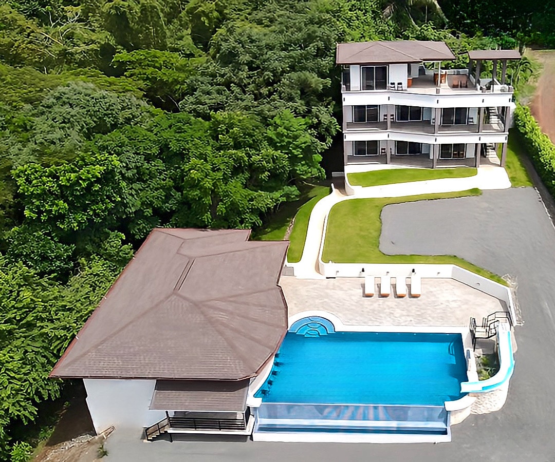 The property features a modern, multi-level building surrounded by lush green foliage. An inviting infinity pool with turquoise water is situated adjacent to the building, complemented by a patio area. A paved driveway leads up to the entrance, enhancing accessibility.