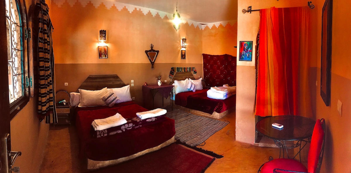 The spacious room features two double beds with decorative red bedding and a cozy atmosphere. A seating area with a small table and chairs is present, accompanied by wall art and colorful fabrics that add cultural charm. Natural light enters through the window, enhancing the inviting space.