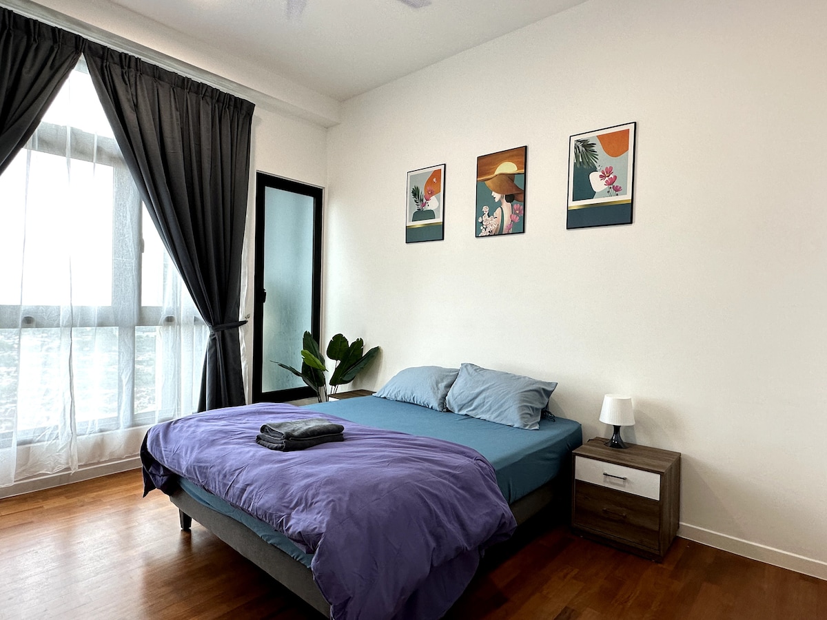 A comfortable bedroom features a spacious bed dressed in blue linens, accompanied by a gray blanket. Two framed artworks adorn the wall above the bed. Natural light filters in through large windows, with sheer curtains softly draping to the side. A small bedside table holds a lamp.