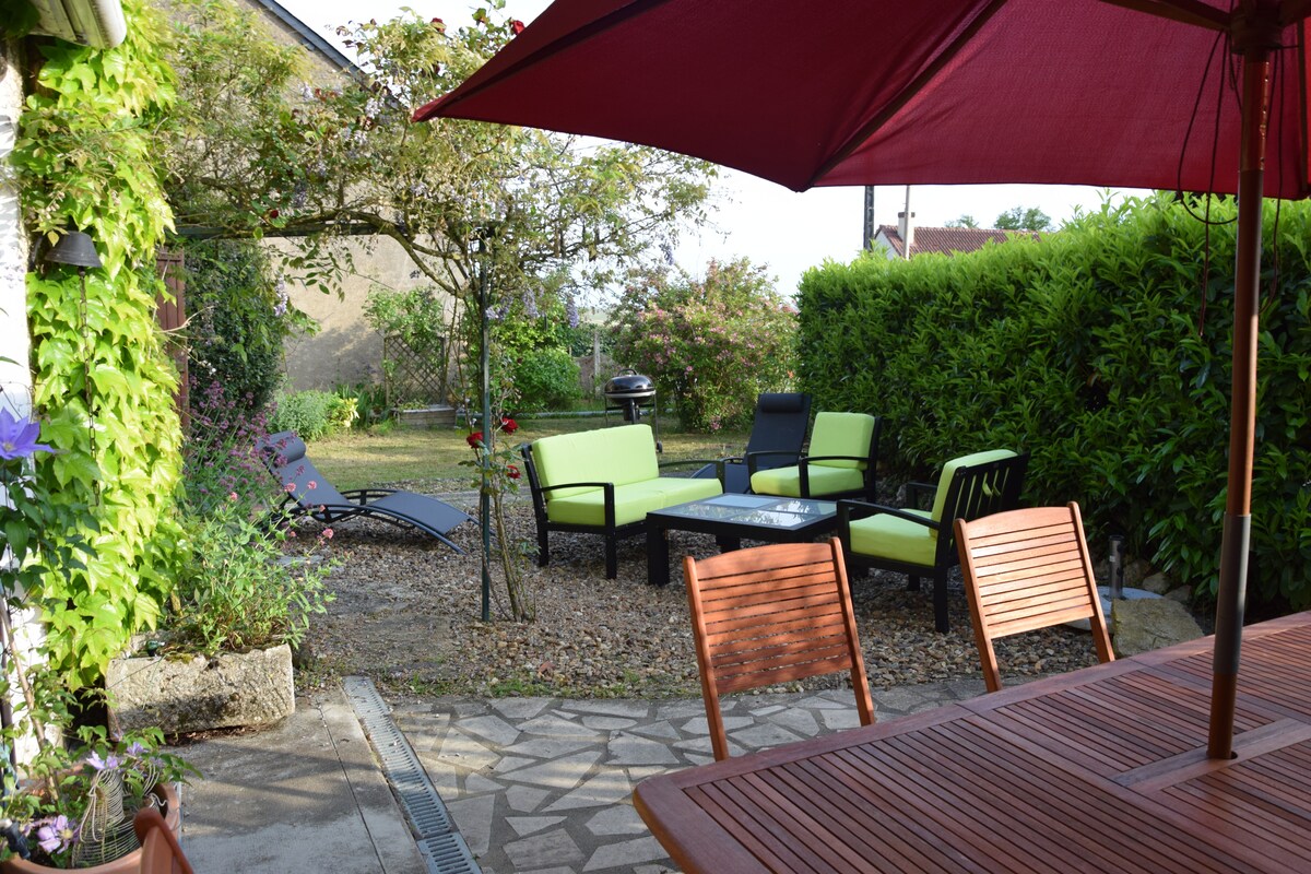 A small garden area is visible, featuring a patio set with green seating and a central table. A lounge chair is positioned nearby, surrounded by greenery and flowering plants. A shaded area is provided by a large umbrella, enhancing the outdoor space.